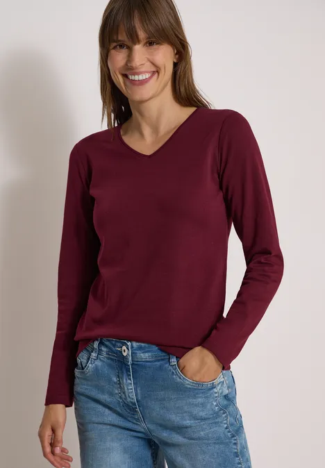 Langarm Shirt in Unifarbe mit V-Neck red wine Frau mit langärmligem burgunderfarbenem V-Ausschnittpullover und blauen Jeans, lächelnd vor neutralem Hintergrund.