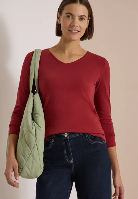 Eine Frau trägt ein rotes Langarmshirt, dunkle Jeans und eine grüne gesteppte Tasche.