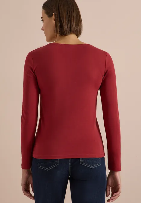 Frau von hinten, trägt rotes Langarmshirt und Jeans vor neutralem Hintergrund