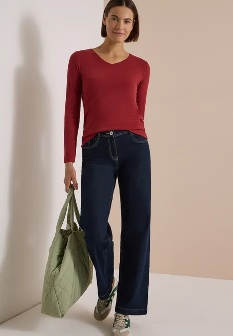 Frau in rotem Langarmshirt, weitem Jeans und grauen Sneakers hält grüne Umhängetasche.