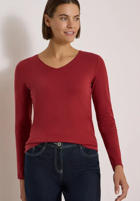 Frau in rotem Langarmshirt und dunklen Jeans posiert vor hellem Hintergrund.