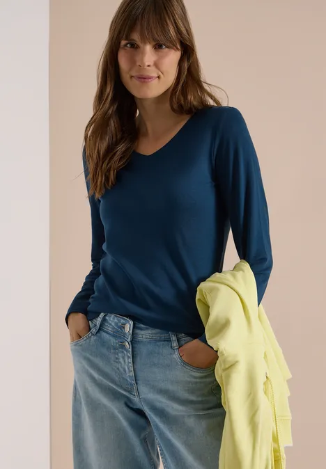 Frau in blauem V-Ausschnitt-Pullover und Jeans hält eine gelbe Jacke und lächelt in die Kamera.