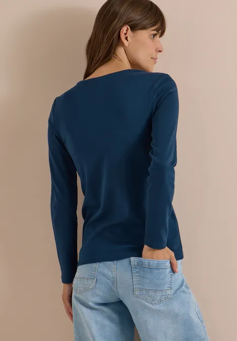 Frau von hinten in dunkelblauem Langarmshirt und hellen Jeans vor beigem Hintergrund.
