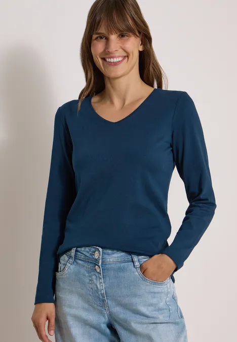 Frau mit blauem Langarmshirt und Jeans lächelt vor hellem Hintergrund.
