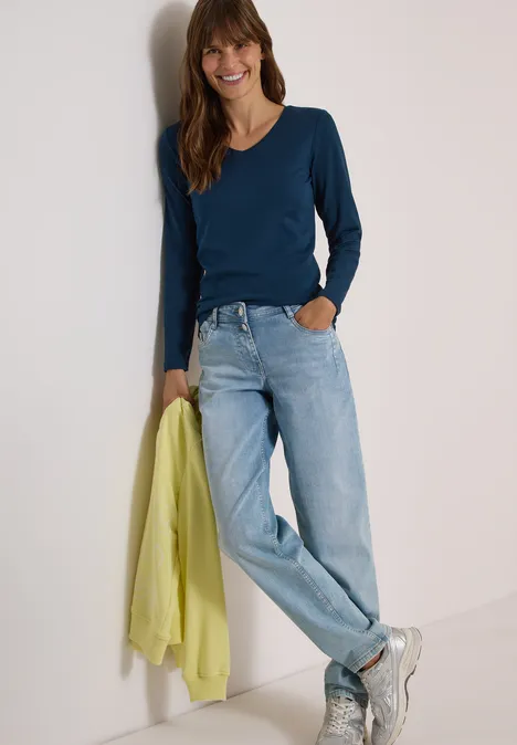 Frau lehnt an Wand, trägt blaues Langarmshirt, helle Jeans und hält einen gelb-grünen Pullover.
