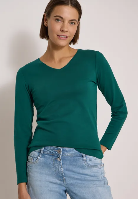 Frau mit kurzem braunem Haar trägt grünes Langarmshirt und helle Jeans, lächelt in die Kamera.
