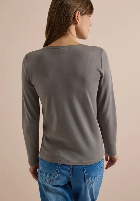 Frau von hinten in grauem Langarmshirt und blauen Jeans vor neutralem Hintergrund.