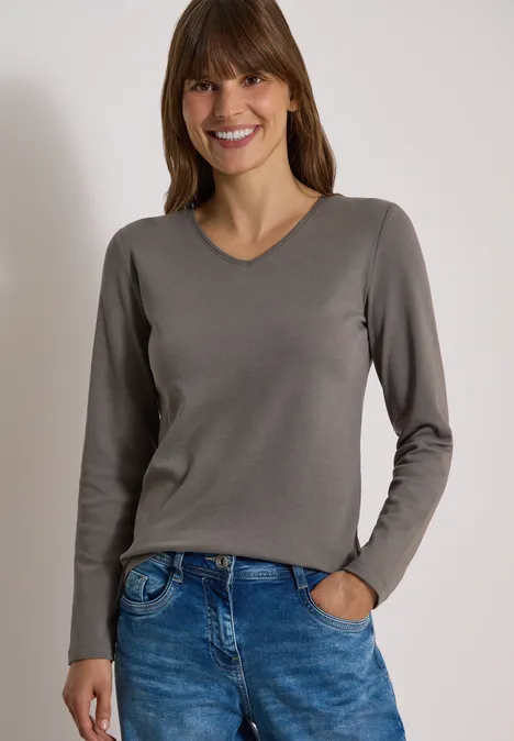 Lächelnde Frau in grauem V-Pullover und blauen Jeans, eine Hand lässig in der Hosentasche.