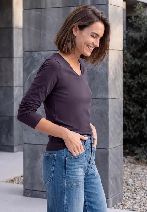 Une femme souriante en jean et haut violet foncé appuyée contre un mur en pierre.