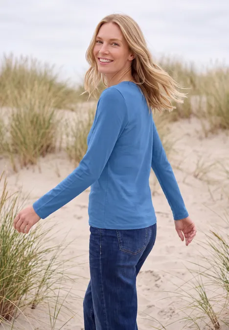 Une femme souriante en pull bleu et jean se tient dans des dunes avec herbes de plage.