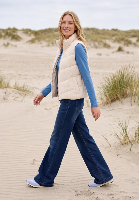 Une femme en gilet, jean et baskets se promène sur les dunes de sable au bord de la mer.