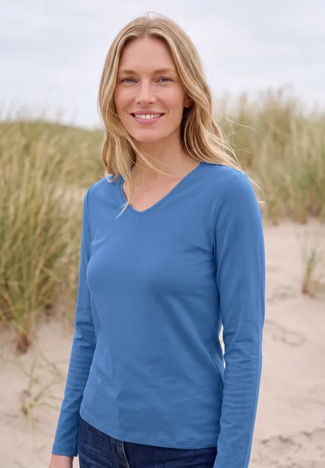 Une femme blonde en t-shirt à manches longues bleu sourit sur une plage devant des herbes de dune.