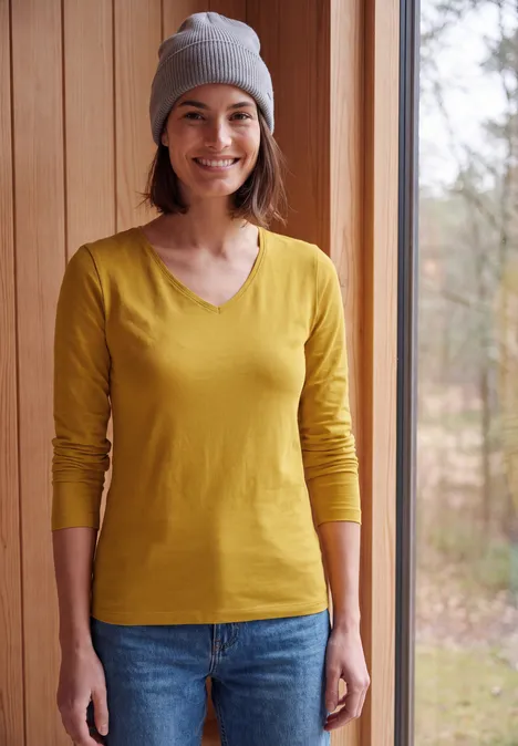 Jeune femme portant un t-shirt jaune à manches longues, un jean bleu et un bonnet gris, souriante dans l’entrée.