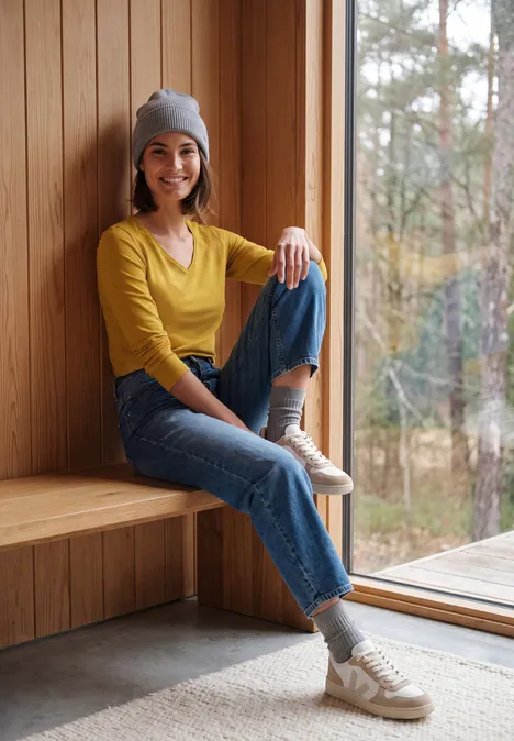 Jeune femme en bonnet et jeans assise près de la fenêtre, regard vers la nature.