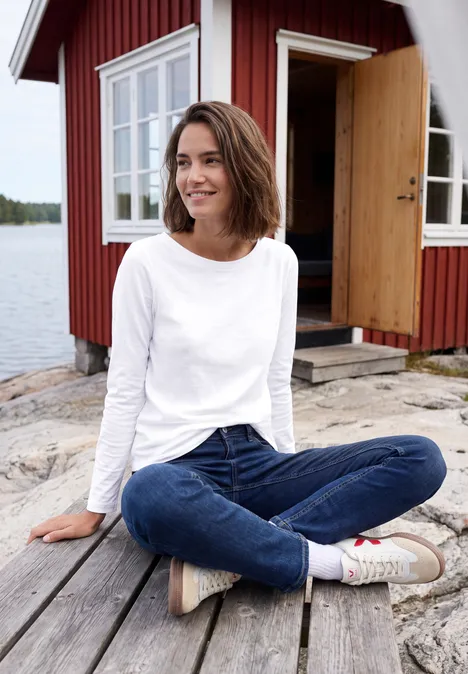Een vrouw zit glimlachend in lotushouding op een houten steiger voor een rood huis aan het water.
