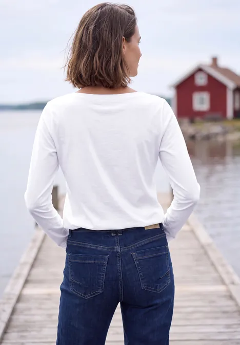 Persoon in wit langarmshirt en spijkerbroek op houten steiger, uitzicht op water en rood huis.