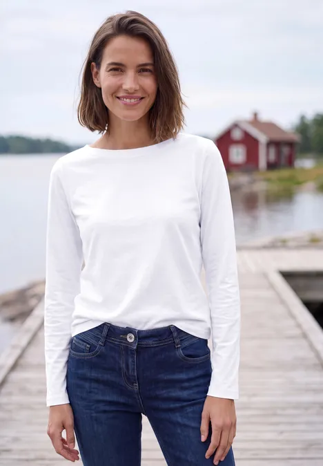 Jonge vrouw in wit shirt en spijkerbroek lacht op een steiger bij een meer met een rood boothuis op de achtergrond.