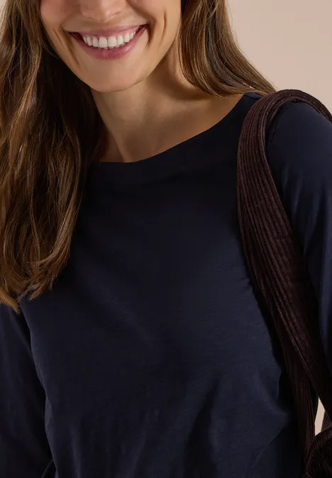 Langarm Shirt mit U-Boot-Ausschnitt urban dark blue Junge Frau mit braunen Haaren lächelt, trägt dunkelblaues Oberteil und braune Schultertasche vor hellem Hintergrund.