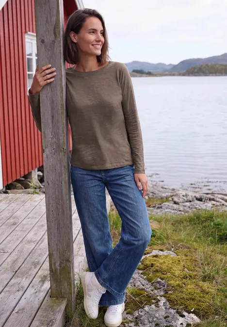 Frau lehnt lächelnd an einem Holzpfosten am Seeufer, trägt Pullover und Jeans.
