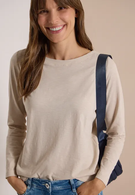 Langarm Shirt mit U-Boot-Ausschnitt cashmere beige Junge Frau lächelt, trägt beige Langarmshirt und Rucksack, steht vor hellem Hintergrund.