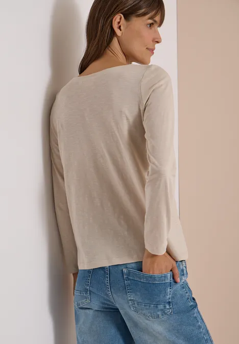 Langarm Shirt mit U-Boot-Ausschnitt cashmere beige Eine Frau in hellbeigem Langarmshirt und blauen Jeans lehnt von hinten an einer hellen Wand.
