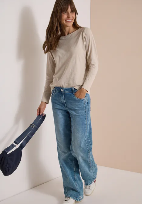 Langarm Shirt mit U-Boot-Ausschnitt cashmere beige Frau in beigem Langarmshirt, weiten Jeans und weißen Sneakern mit blauer Tasche steht lächelnd in Raumecke.