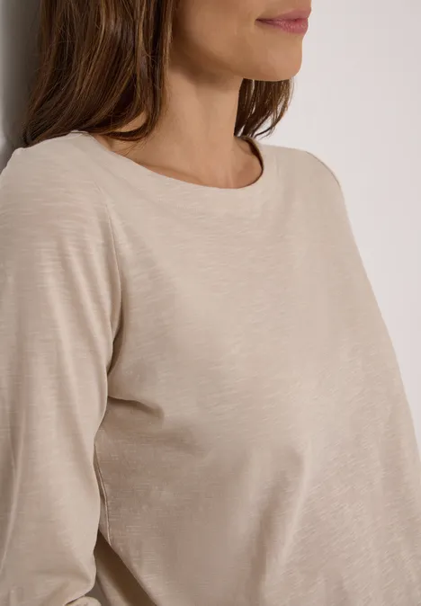 Langarm Shirt mit U-Boot-Ausschnitt cashmere beige Frau mit beigem Langarmshirt lehnt sich an eine weiße Wand.