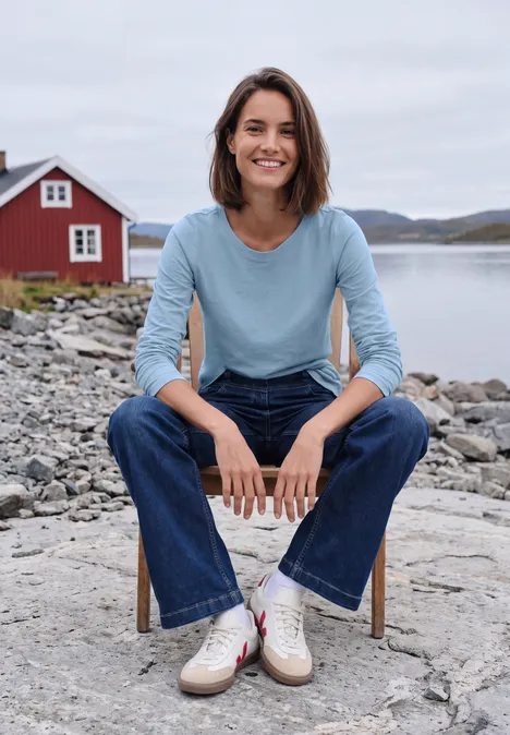 Eine lächelnde Frau in Jeans und blauem Shirt sitzt auf einem Stuhl am felsigen Ufer mit rotem Haus im Hintergrund.
