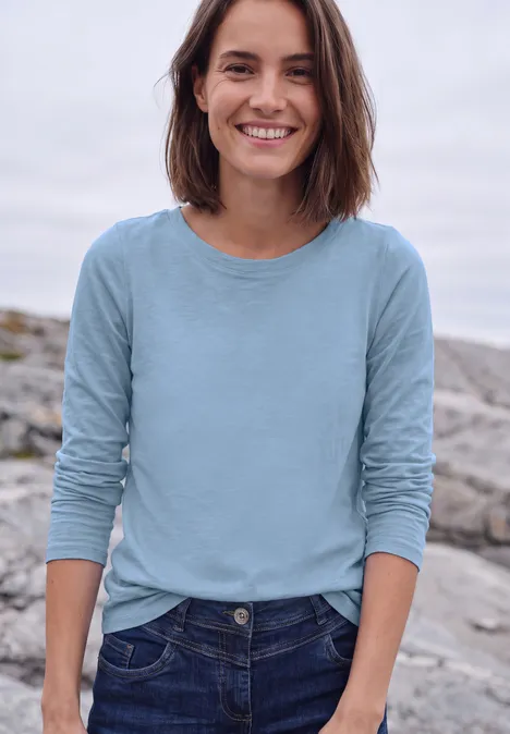 Eine lächelnde Frau in Jeans und hellblauem Langarmshirt steht auf Felsen am Wasser.