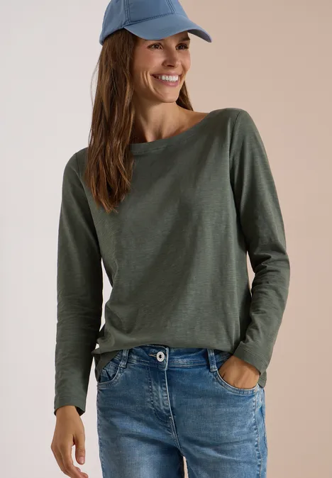 Langarm Shirt mit U-Boot-Ausschnitt dried olive green Frau trägt grünes Longsleeve, Jeans und blauen Cap, lächelt.
