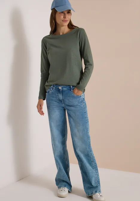 Langarm Shirt mit U-Boot-Ausschnitt dried olive green Frau in weitem Jeans, grünem Langarmshirt, weißen Sneakern und blauer Baseballkappe vor hellem Hintergrund.