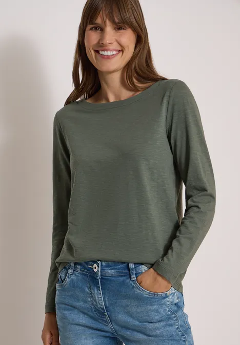 Langarm Shirt mit U-Boot-Ausschnitt dried olive green Eine lächelnde Frau in grüner Langarmshirt und blauen Jeans vor hellem Hintergrund.