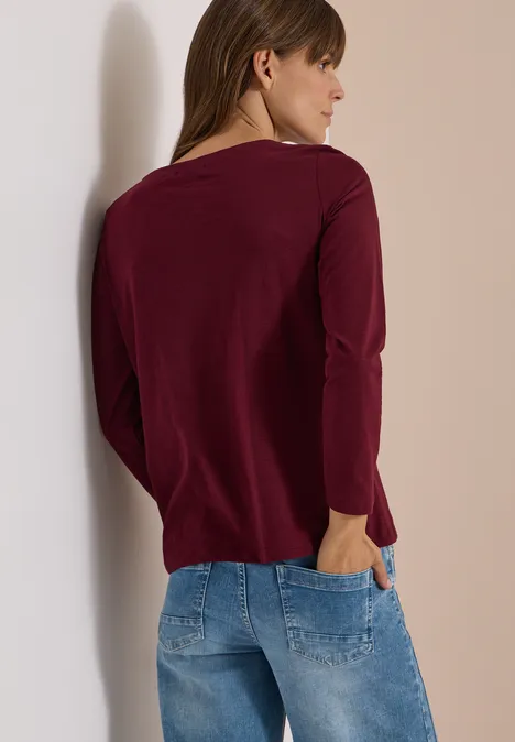 Langarm Shirt mit U-Boot-Ausschnitt red wine Eine Frau lehnt an einer Wand, trägt einen burgunderfarbenen Langarmpullover und Jeans und steht seitlich zur Kamera.