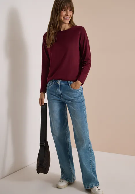 Langarm Shirt mit U-Boot-Ausschnitt red wine Frau in burgunderrotem Pullover, weitem blauen Jeans und schwarzer Tasche steht vor neutralem Hintergrund.