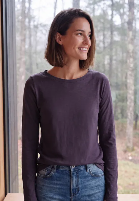 Eine lächelnde Frau mit braunem Bob, lila Longsleeve und Jeans steht am Fenster vor Wald