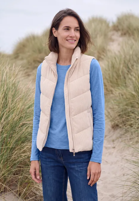 T-shirt à manches longues et col bateau north blue Eine Frau in beige Weste und blauem Langarmshirt steht lächelnd am Strand vor Dünen.