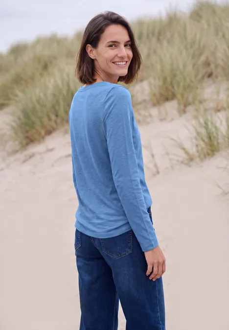 T-shirt à manches longues et col bateau north blue Eine Frau in blauem Langarmshirt und Jeans steht lächelnd auf Dünenstrand, blickt über die Schulter.