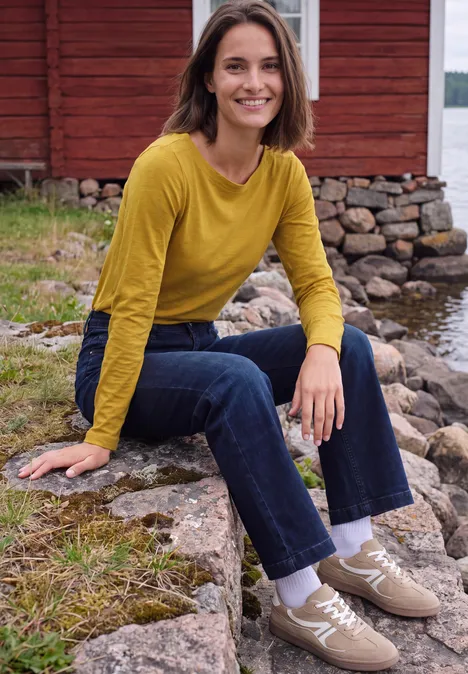 T-shirt à manches longues et col bateau golden honey Frau in gelbem Pullover und Jeans sitzt auf Steinen vor einem roten Holzhaus am Wasser.