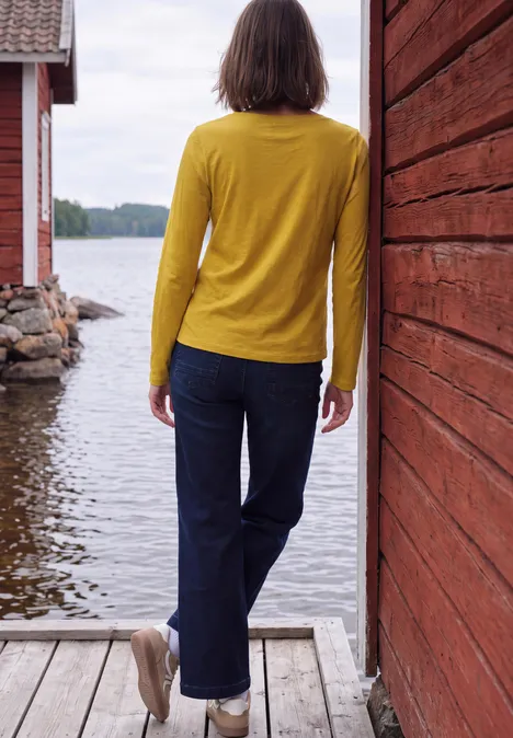 T-shirt à manches longues et col bateau golden honey Frau im gelben Pullover steht rückwärts auf Steg zwischen roten Holzhäusern und blickt auf den See.