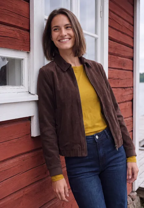 T-shirt à manches longues et col bateau golden honey Eine lächelnde Frau in brauner Jacke und gelbem Shirt lehnt an einer rot gestrichenen Holzwand.
