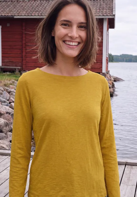 T-shirt à manches longues et col bateau golden honey Eine lächelnde Frau in senfgelbem Top steht an einem Steg vor einem roten Haus am See.