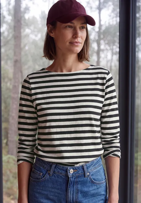 Langarm Shirt mit Boat Neck und Streifen Black Frau mit roter Baseballmütze, gestreiftem Langarmshirt und Jeans steht vor einem Fenster mit Waldblick.