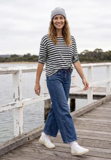 Une femme en jean, marinière, bonnet et baskets marche en souriant sur une jetée au bord de l’eau.