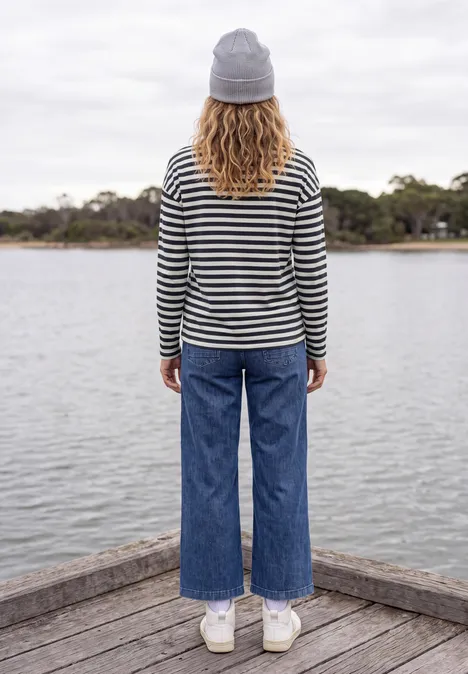 Personne en pull rayé, jean et bonnet, debout sur une jetée, contemplant un lac paisible.