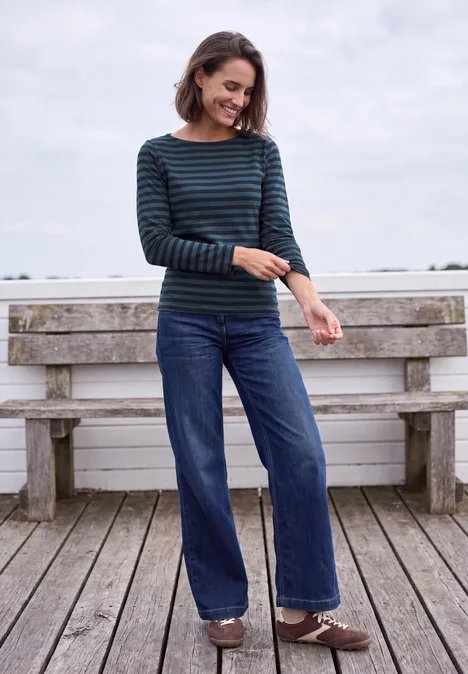 Langarm Shirt mit Boat Neck und Streifen urban dark blue Frau steht auf Holzdeck vor Bank, trägt gestreiftes Oberteil, weite Jeans und lächelt entspannt.