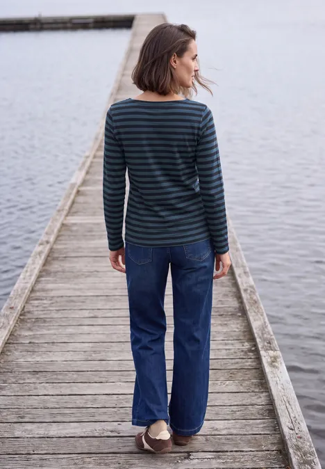 Langarm Shirt mit Boat Neck und Streifen urban dark blue Frau in gestreiftem Pullover und Jeans steht auf einem Holzsteg am Wasser, Blick nach links.