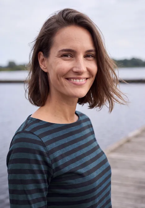Langarm Shirt mit Boat Neck und Streifen urban dark blue Eine lächelnde Frau mit kurzen braunen Haaren und gestreiftem Oberteil steht auf einem Steg am Wasser.