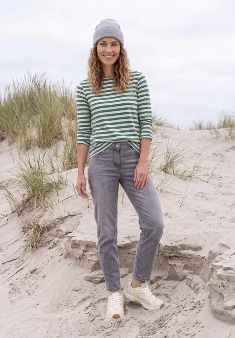 Lächelnde Frau mit grüner Streifenbluse, grauen Jeans und Mütze steht auf sandigen Dünen.