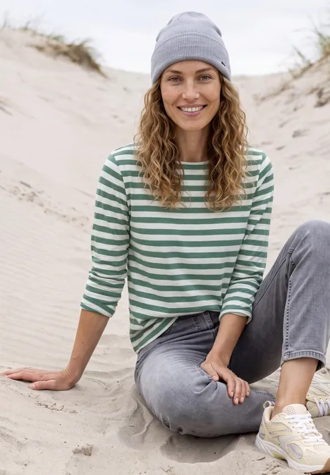 Frau sitzt lächelnd auf Sanddüne, trägt Ringelshirt, Jeans, Mütze und Sneaker.