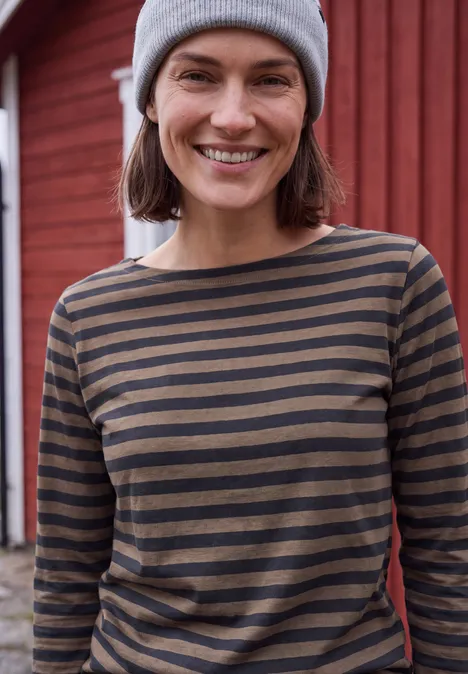 Une femme souriante en bonnet gris et t-shirt à rayures devant un mur de bois rouge.